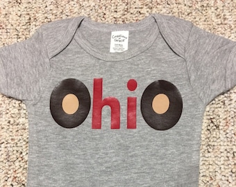 May include: Gray baby onesie with the word "Ohio" in red letters and two brown circles with tan centers.