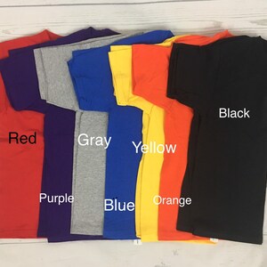May include: A stack of eight t-shirts in different colours, including red, purple, grey, blue, yellow, orange, and black. The shirts are folded and stacked on a white wooden surface.