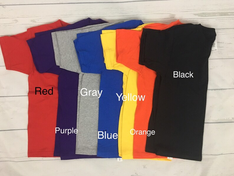 May include: A stack of eight t-shirts in different colours, including red, purple, grey, blue, yellow, orange, and black. The shirts are folded and stacked on a white wooden surface.