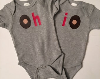 May include: Two gray baby onesies with the word "Ohio" printed in red letters. Each onesie has a black and brown circle with a smaller brown circle inside, representing a record.