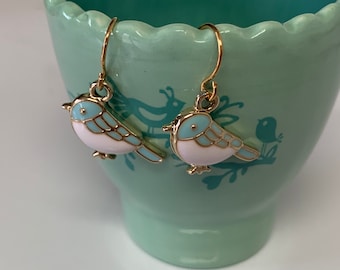 May include: A pair of gold-coloured bird earrings. Each earring features a light blue and white enamel bird charm with gold accents. The earrings are suspended from gold-coloured ear wires, showcasing a charming and whimsical design.