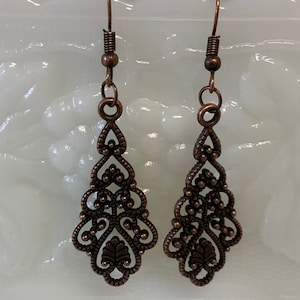May include: A pair of copper filigree dangle earrings with a teardrop shape. The earrings have a delicate, intricate design with small beads and swirls.
