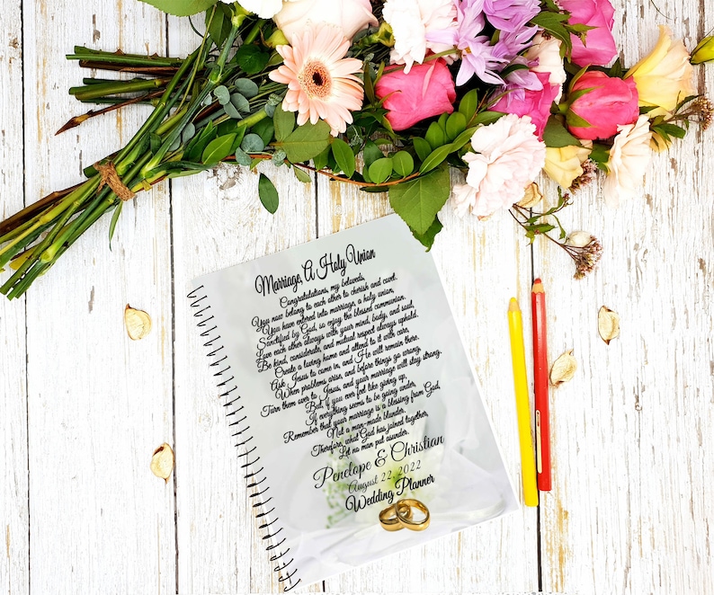 May include: A white spiral bound wedding planner with a floral design and the text "Marriage A Holy Union" and "Penelope & Christian August 22, 2022 Wedding Planner".
