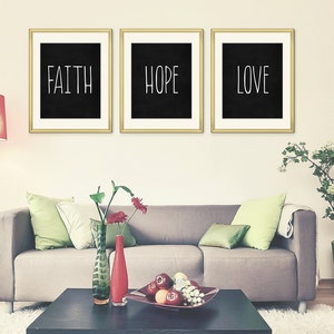 May include: Three framed black and white prints with gold frames hanging on a white wall. The prints have the words "Faith", "Hope", and "Love" written in white on a black background.