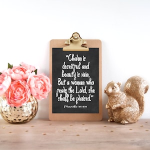 May include: A black chalkboard with a white inscription on a wooden clipboard. The inscription reads "Charm is deceitful and beauty is vain, But a woman who fears the Lord, she shall be praised."  Proverbs 31:30. The clipboard is on a wooden surface with a gold vase of pink flowers and a gold squirrel figurine.