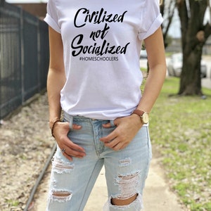 May include: A white t-shirt with the text "Civilized not Socialized" and the hashtag "#HOMESCHOOLERS" printed in black. The person wearing the shirt is standing on a sidewalk with a green lawn in the background.