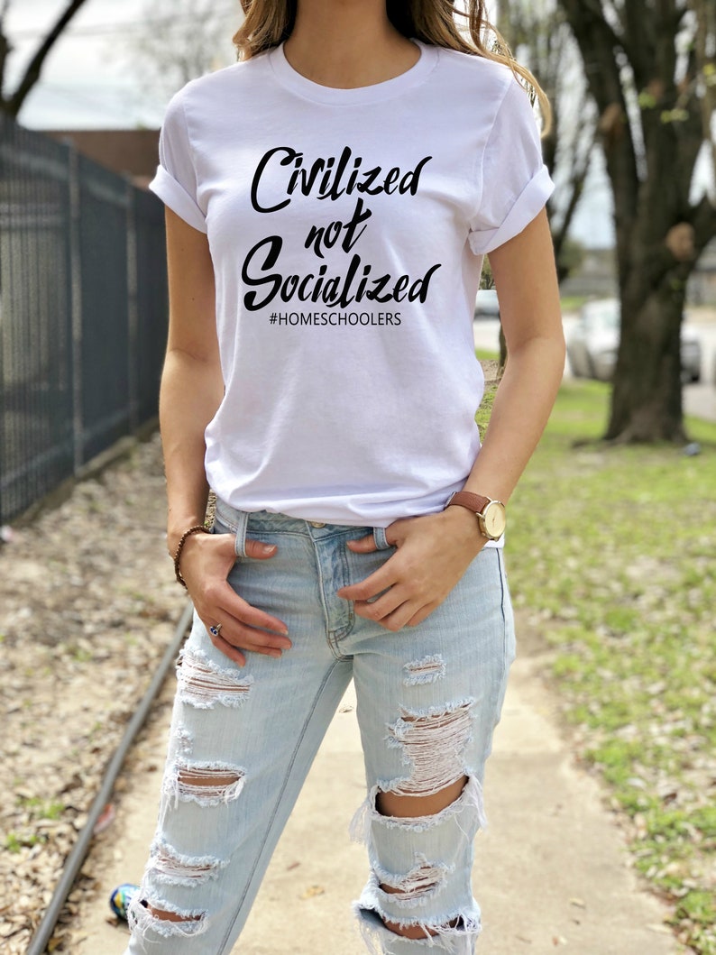 May include: A white t-shirt with the text "Civilized not Socialized" and the hashtag "#HOMESCHOOLERS" printed in black. The person wearing the shirt is standing on a sidewalk with a green lawn in the background.