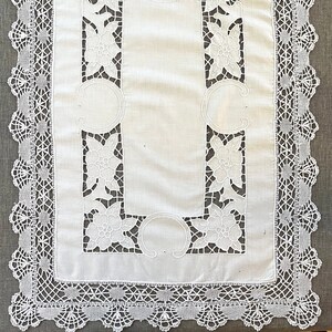 May include: White rectangular tablecloth with a floral design and a decorative lace border. The centre features cut-out floral patterns and curved accents. The intricate lace detailing adds a delicate touch to this elegant table linen.