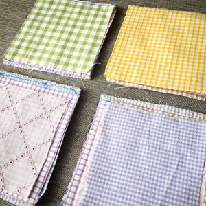 以下が含まれることがあります： 異なる色と模様の布の正方形が4つの積み重ねになっています。一番上の積み重ねは緑と白のギンガムチェック、2番目の積み重ねは黄と白のギンガムチェック、3番目の積み重ねは白地に薄いピンクの菱形模様、4番目の積み重ねは薄い紫と白のギンガムチェックです。