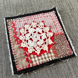 May include: A square patchwork quilt block with a red, white and brown colour scheme. The centre of the block features a white crocheted doily on a red background.