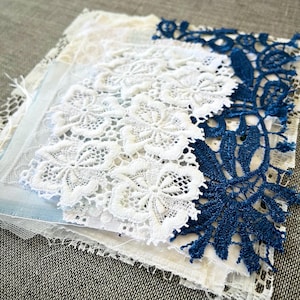 May include: A stack of white and blue lace fabric scraps. The lace is delicate and intricate, with a floral pattern. The fabric is perfect for crafting and sewing projects.