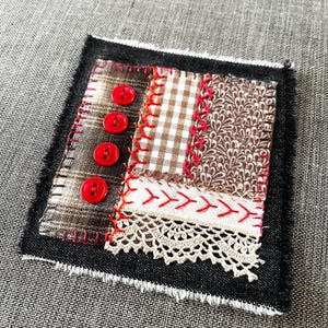 May include: A square fabric patch featuring a black denim base with frayed edges. The surface is decorated with four red buttons, a brown plaid section, a brown and white gingham square, a brown floral print, and a piece of cream-coloured lace.