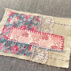 May include: A patchwork textile featuring floral fabric in pink and blue, with white lace and red stitching. The piece is sewn onto a beige linen background with blue and purple stitching.