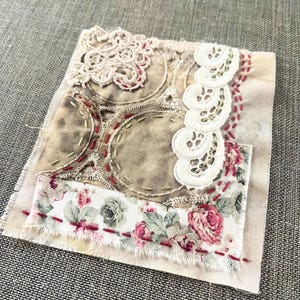 May include: A handmade textile art piece with layered fabrics. The design includes a floral print, lace, and stitched details in beige, brown, and red. The piece has a vintage, handcrafted aesthetic.