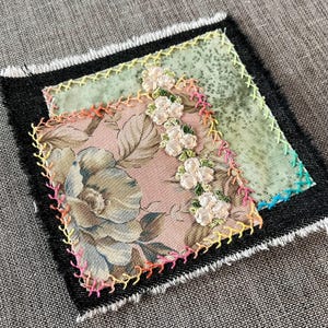 May include: A small square patchwork quilt with a floral print fabric in the centre. The quilt is edged with black fabric and has colourful stitching around the edges.