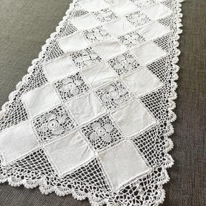 May include: A white rectangular table runner featuring a repeating pattern of solid fabric squares alternating with intricate crocheted floral squares. The runner is finished with a delicate scalloped crochet lace border along all four edges.