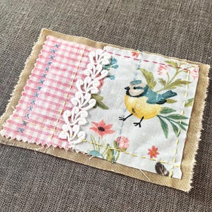 May include: A rectangular fabric patch featuring a pink and white gingham section joined to a light blue floral print section with a bird illustration. A white lace trim separates the fabrics, and yellow and blue hand-stitched details are visible.