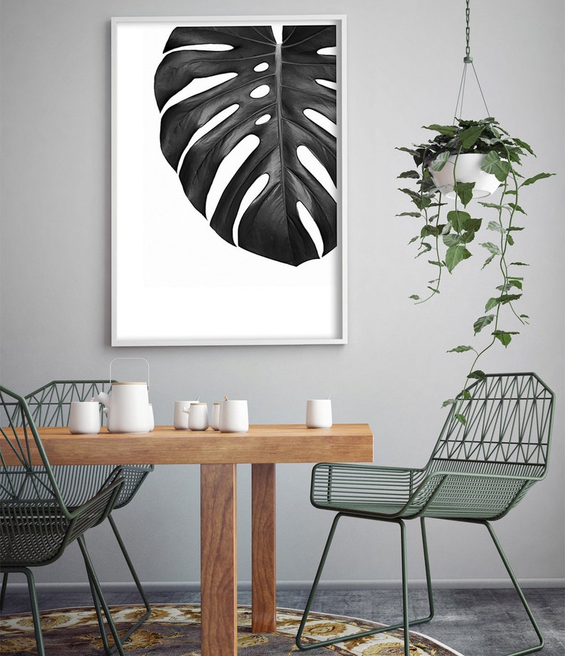 May include: A black and white framed print of a large tropical leaf, hanging on a gray wall above a wooden table with two green metal chairs. A hanging plant with green leaves is in the background.