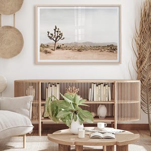 May include: A framed landscape print featuring a Joshua tree in a desert setting. The print is in a light wood frame and hangs above a wooden cabinet with books and decor. The room has a neutral color palette with natural textures.