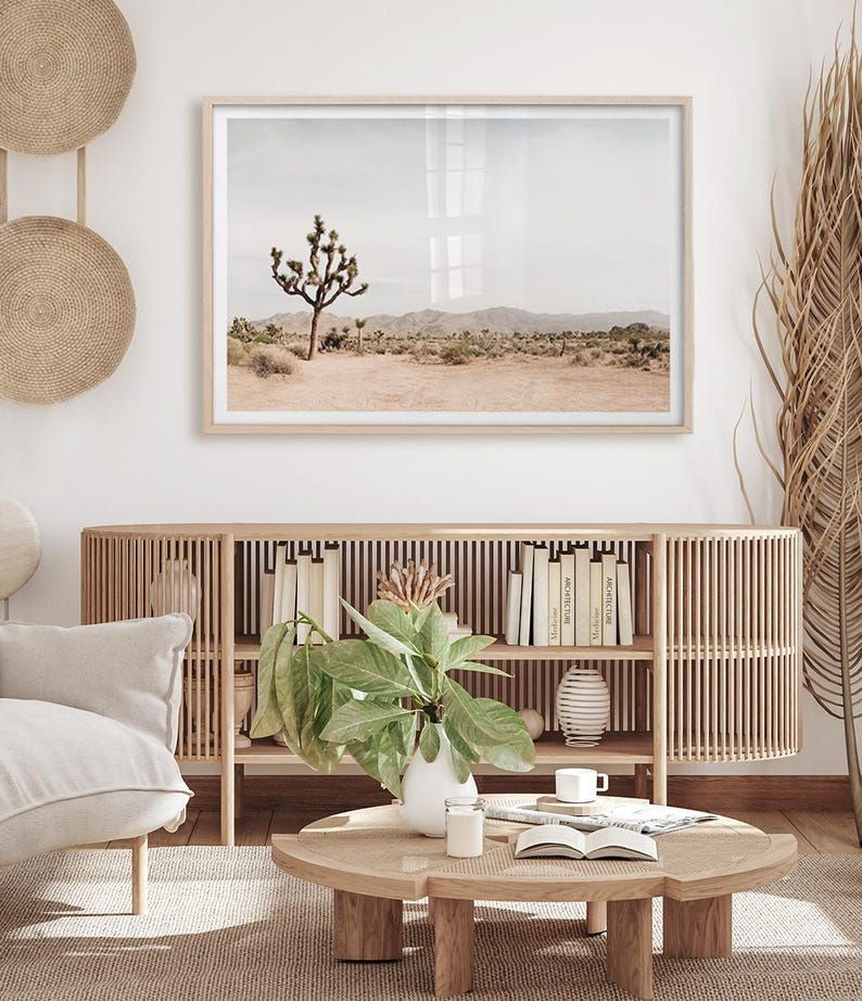 May include: A framed landscape print featuring a Joshua tree in a desert setting. The print is in a light wood frame and hangs above a wooden cabinet with books and decor. The room has a neutral color palette with natural textures.