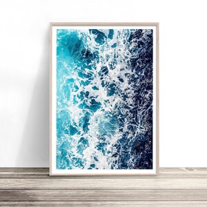 May include: A rectangular wall art print in a light wood frame featuring an aerial view of ocean waves. The image displays a gradient of deep navy blue water transitioning into bright turquoise, with white sea foam crashing across the surface.