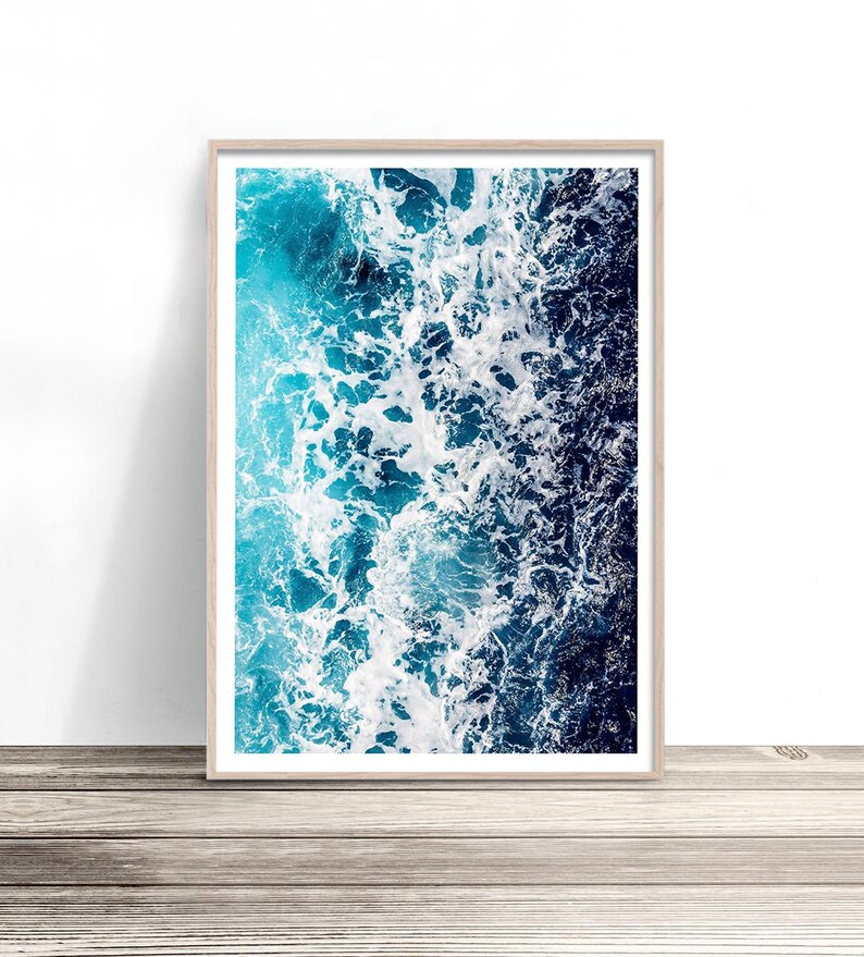 May include: A rectangular wall art print in a light wood frame featuring an aerial view of ocean waves. The image displays a gradient of deep navy blue water transitioning into bright turquoise, with white sea foam crashing across the surface.