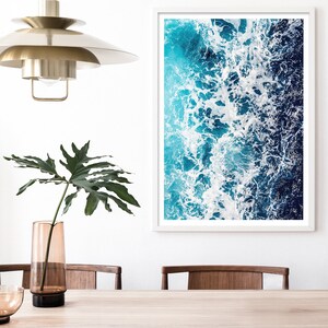 May include: A framed wall art print featuring an aerial view of ocean waves with white sea foam, deep blue and turquoise water. The artwork is displayed on a white wall next to a tall amber glass vase with a green leaf and a brass pendant light.