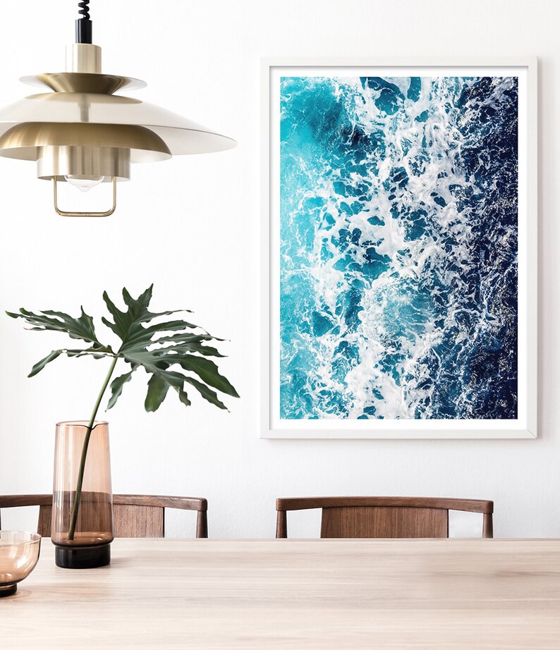May include: A framed wall art print featuring an aerial view of ocean waves with white sea foam, deep blue and turquoise water. The artwork is displayed on a white wall next to a tall amber glass vase with a green leaf and a brass pendant light.
