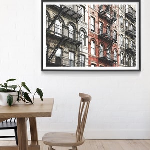May include: A framed photograph of a row of brick buildings with fire escapes. The buildings are painted in shades of gray and red. The fire escapes are made of black metal.