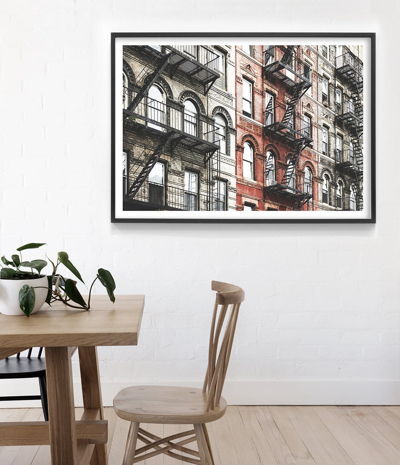 May include: A framed photograph of a row of brick buildings with fire escapes. The buildings are painted in shades of gray and red. The fire escapes are made of black metal.