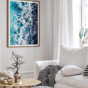 May include: A rectangular wall art print featuring an aerial view of ocean waves with deep blue and turquoise water contrasted by white sea foam, enclosed in a thin wooden frame and displayed on a light-colored wall above a white sofa.
