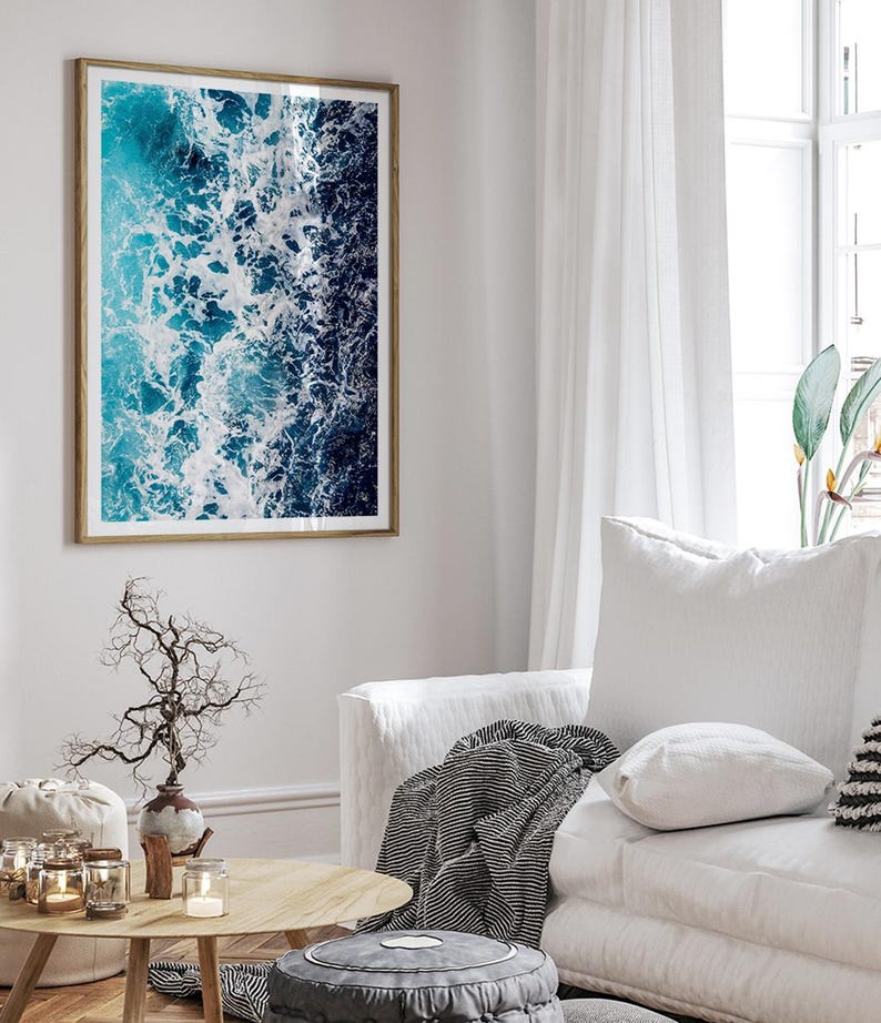 May include: A rectangular wall art print featuring an aerial view of ocean waves with deep blue and turquoise water contrasted by white sea foam, enclosed in a thin wooden frame and displayed on a light-colored wall above a white sofa.