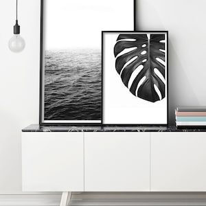 May include: Two black and white framed prints on a white cabinet with a black marble top. The print on the left shows a black and white image of the ocean. The print on the right shows a black and white image of a monstera leaf.