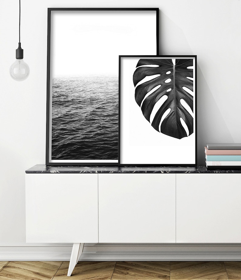May include: Two black and white framed prints on a white cabinet with a black marble top. The print on the left shows a black and white image of the ocean. The print on the right shows a black and white image of a monstera leaf.
