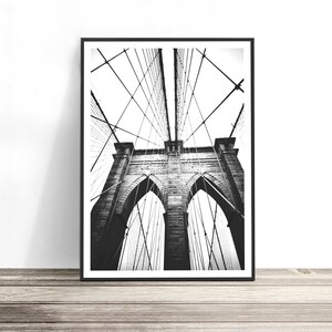 May include: A black and white framed wall art print featuring a low-angle perspective of the Brooklyn Bridge. The image captures the stone towers with arched openings and the intricate network of steel suspension cables reaching upward.