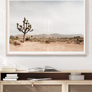 May include: A framed landscape print featuring a Joshua tree in a desert environment. The image showcases a muted color palette with a beige foreground, a Joshua tree, and mountains in the background. The print is in a light wood frame.