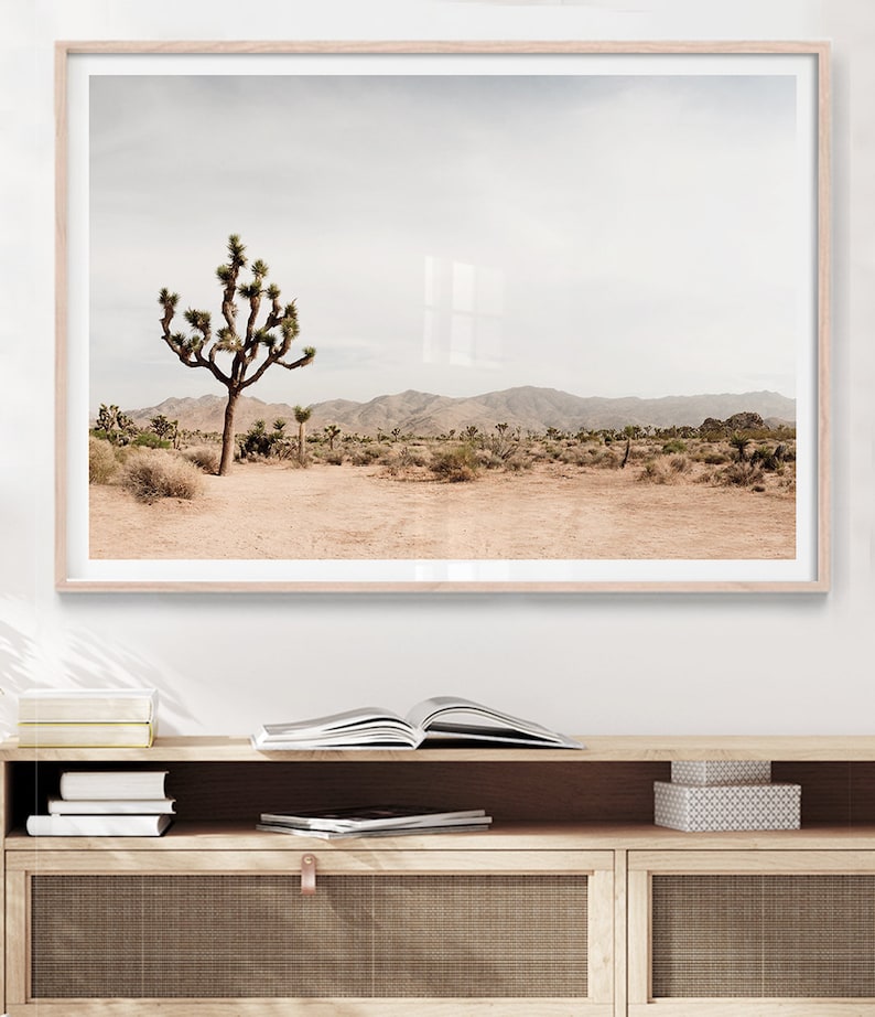 May include: A framed landscape print featuring a Joshua tree in a desert environment. The image showcases a muted color palette with a beige foreground, a Joshua tree, and mountains in the background. The print is in a light wood frame.