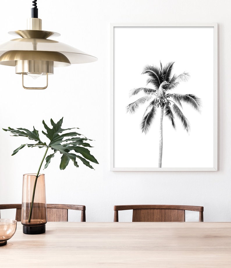 May include: Black and white photograph of a palm tree in a white frame. The frame is hanging on a white wall above a wooden table with two chairs. A vase with a green plant is on the table.