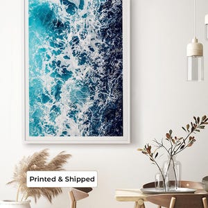 May include: A rectangular wall art print featuring an aerial view of ocean waves crashing against a shoreline. The image displays a gradient of deep navy blue to bright turquoise water with white sea foam patterns encased in a white frame.