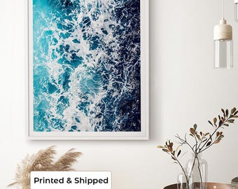May include: A rectangular wall art print featuring an aerial view of ocean waves crashing against a shoreline. The image displays a gradient of deep navy blue to bright turquoise water with white sea foam patterns encased in a white frame.