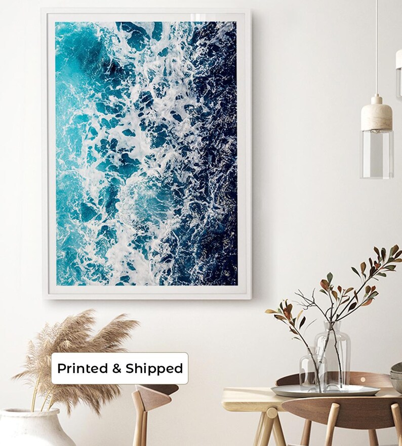 May include: A rectangular wall art print featuring an aerial view of ocean waves crashing against a shoreline. The image displays a gradient of deep navy blue to bright turquoise water with white sea foam patterns encased in a white frame.