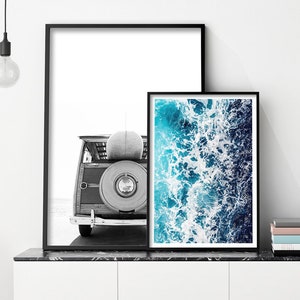 May include: Two framed wall art prints in black frames. The left print is a black and white photograph of the rear of a vintage woody station wagon with a surfboard on top. The right print is a vibrant aerial view of blue ocean waves.
