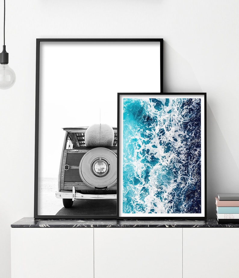 May include: Two framed wall art prints in black frames. The left print is a black and white photograph of the rear of a vintage woody station wagon with a surfboard on top. The right print is a vibrant aerial view of blue ocean waves.