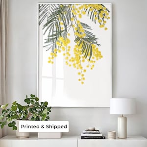 May include: A vertical wall art print in a thin white frame featuring a botanical illustration of bright yellow mimosa flowers and dark green feathery leaves against a clean white background, displayed as a piece of home decor.