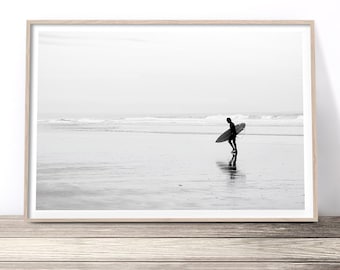 May include: Black and white photograph of a surfer walking along a beach with a surfboard under their arm. The surfer is silhouetted against the bright sky and the water is calm and reflective.