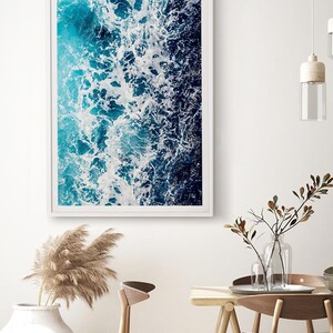 May include: A rectangular wall art print featuring an aerial view of ocean waves crashing against the shore. The image displays a gradient of deep navy blue to bright turquoise water with white sea foam patterns captured in a white frame.