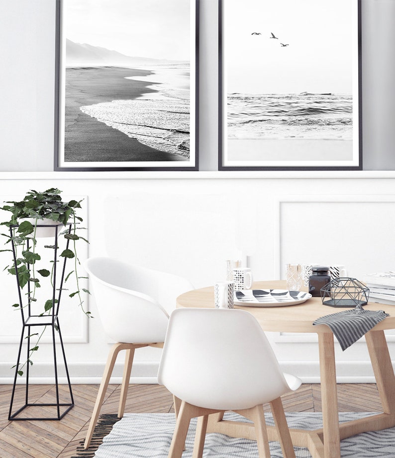 May include: Two black and white framed photographs of a beach scene with waves crashing on the shore. The photographs are hanging on a white wall above a wooden table with two white chairs. A potted plant is on the left side of the image.
