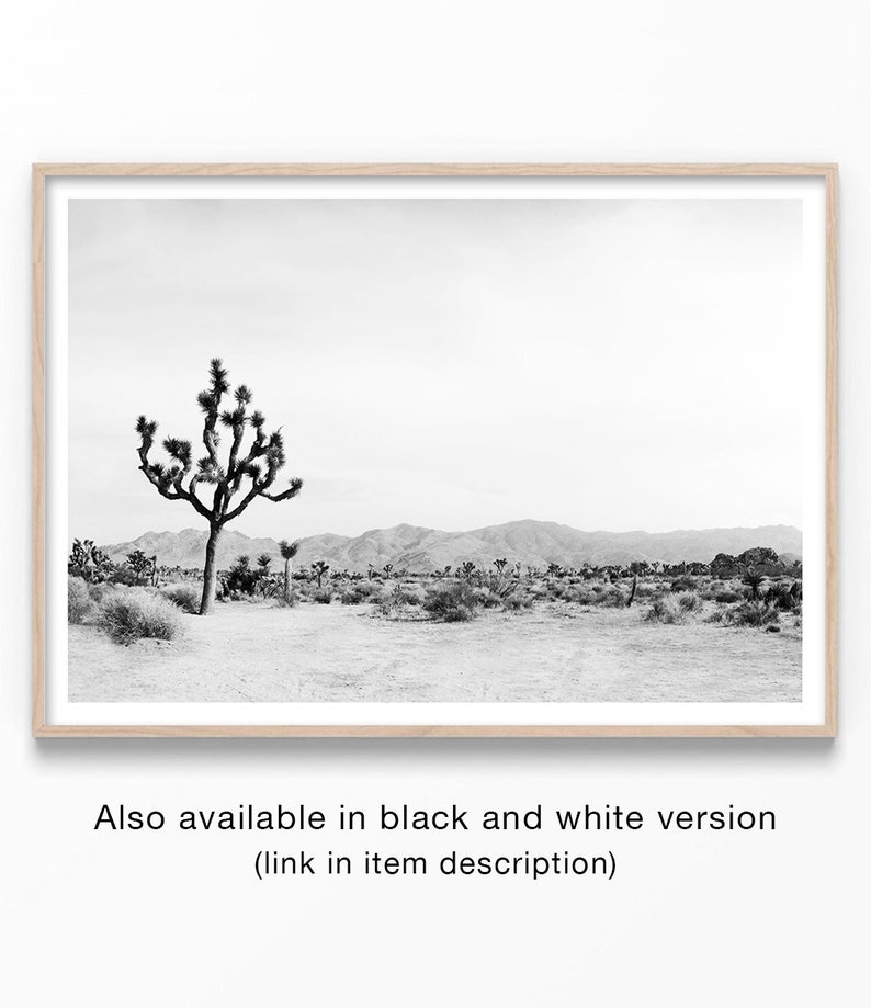 May include: Black and white framed print of a desert landscape featuring a Joshua tree in the foreground. Mountains are visible in the background under a cloudy sky. The print is in a light wood frame. Text at the bottom reads: "Also available in black and white version (link in item description)".