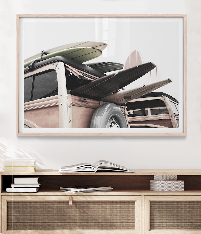 May include: A framed print featuring a vintage wood-paneled station wagon with surfboards strapped to the roof. The surfboards are in various colors and shapes. The print is displayed above a wooden cabinet with books and decorative boxes.