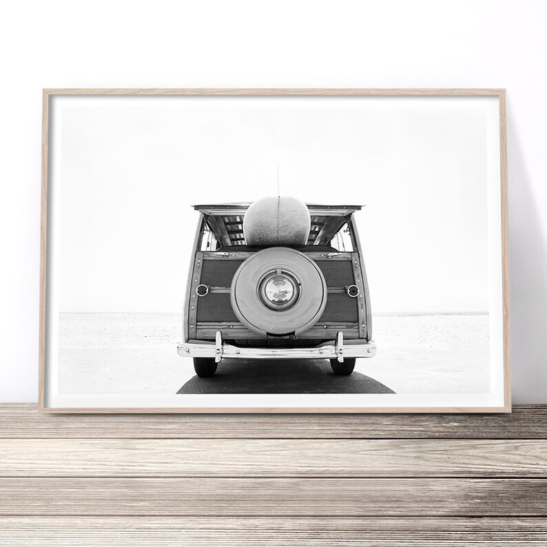 K&ouml;nnte beinhalten: Ein Schwarzwei&szlig;foto eines alten Holz-Kombiwagens mit einem Surfbrett auf dem Dach. Das Auto steht auf einem Strand mit dem Meer im Hintergrund.
